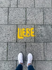 Foto zu LIEBESDINGE: Graue Betonplatten von oben gesehen. Das Wort 'Liebe' steht in Gelb darauf gesprüht. Die Fotografin steht vor dem Wort und fotografiert ihre Füße, die in Turnschuhen vor dem Wort stehen.