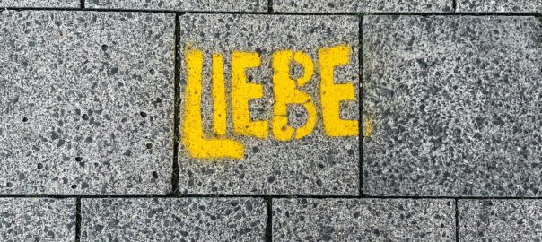Foto zu LIEBESDINGE: Graue Betonplatten von oben gesehen. Das Wort 'Liebe' steht in Gelb darauf gesprüht. Die Fotografin steht vor dem Wort und fotografiert ihre Füße, die in Turnschuhen vor dem Wort stehen.